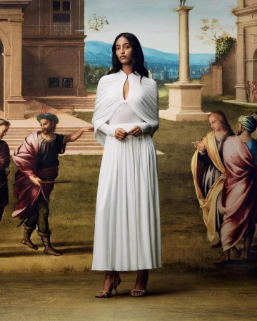 Elegant woman in long white dress poses in front of a painted backdrop with classical architecture and figures, under soft light.