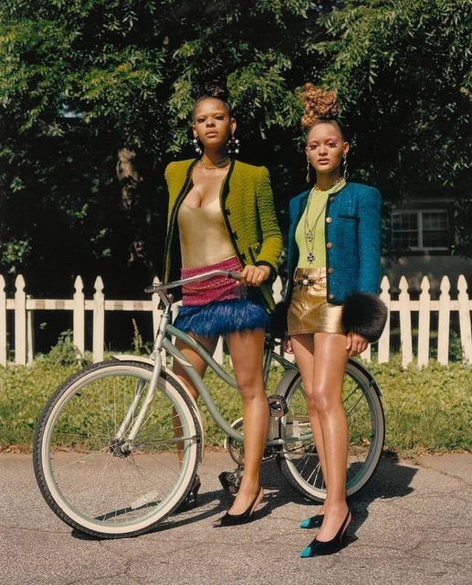 Two adult females posing with a light blue bicycle on an asphalt street against a backdrop of a white picket fence and green trees.
