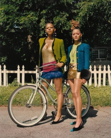 Two adult females posing with a light blue bicycle on an asphalt street against a backdrop of a white picket fence and green trees.