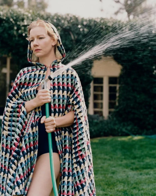 Woman in patterned cape holds green hose spraying water in garden, with green lawn and ivy-covered house in the background.