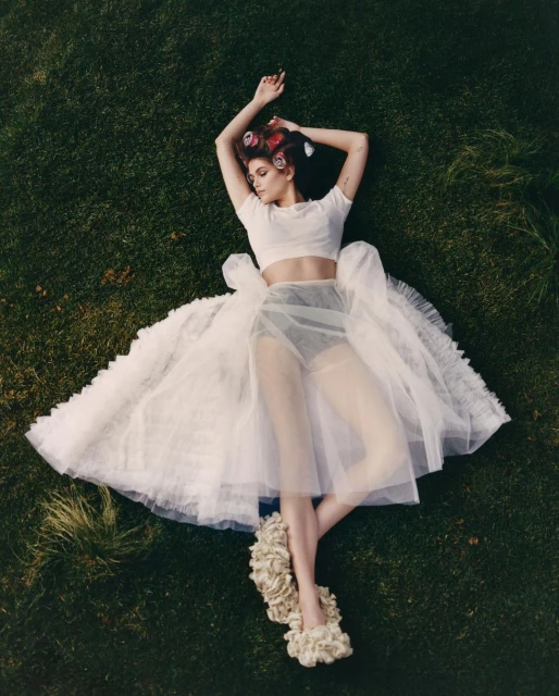 Young woman lies on green grass wearing a white crop top, tulle skirt, and hair curlers, posing for a fashion or beauty editorial.