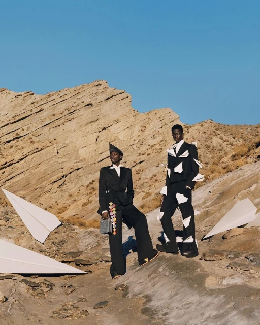 Two African-descent models pose in a desert setting wearing fashion suits with paper airplane details under a clear blue sky.