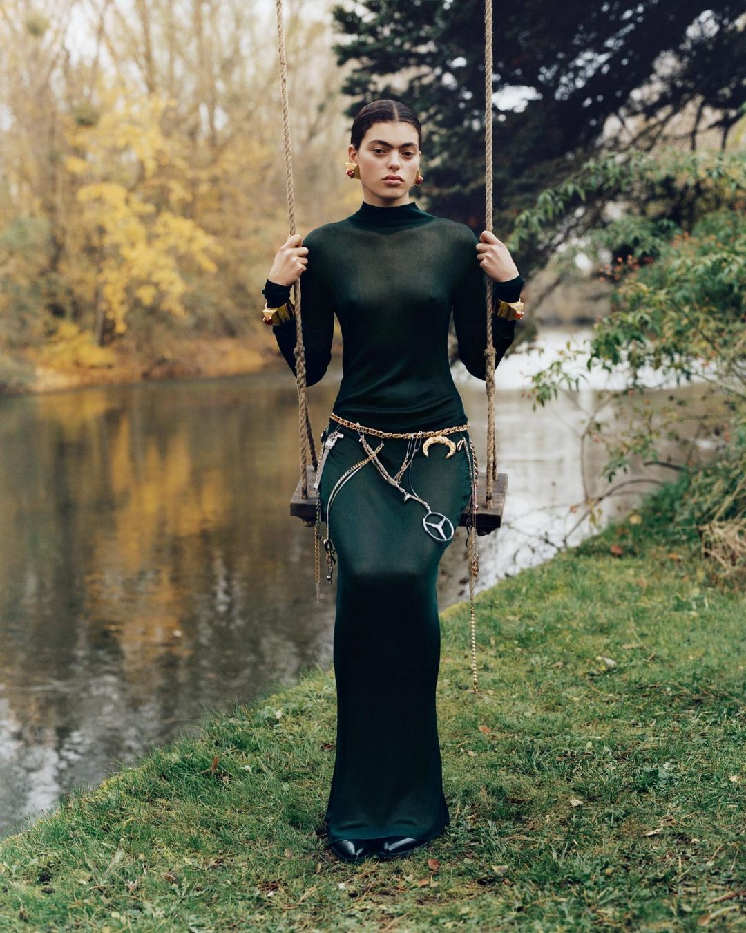 Young woman in a dark green dress and gold jewelry poses on a swing by a river in a wooded area with fall foliage on the trees.