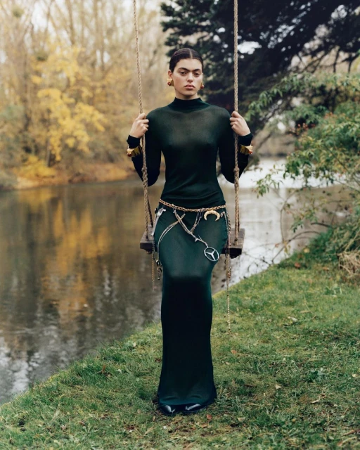 Young woman in a dark green dress and gold jewelry poses on a swing by a river in a wooded area with fall foliage on the trees.