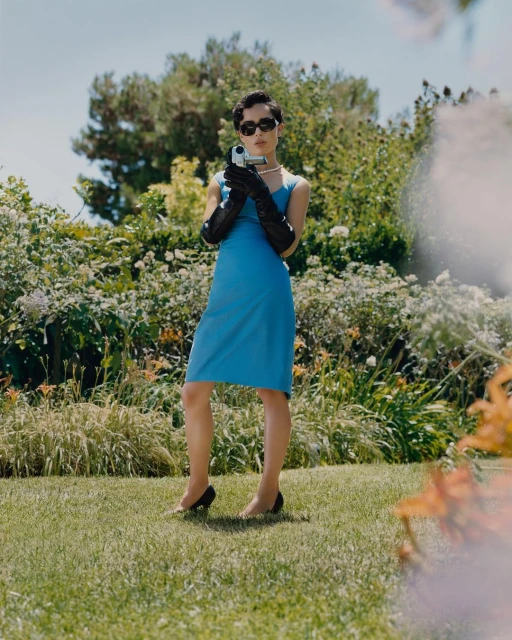 Young woman in blue dress, sunglasses, and gloves holds vintage camcorder outdoors against garden background on a sunny day.