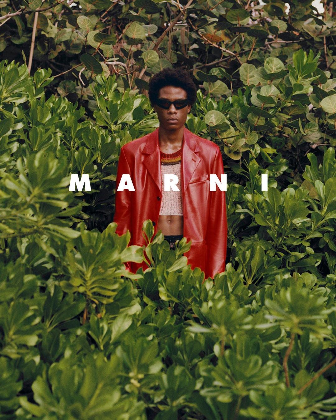 Adult male model in red leather jacket posing in green foliage, outdoor fashion shot with prominent MARNI logo, natural light.