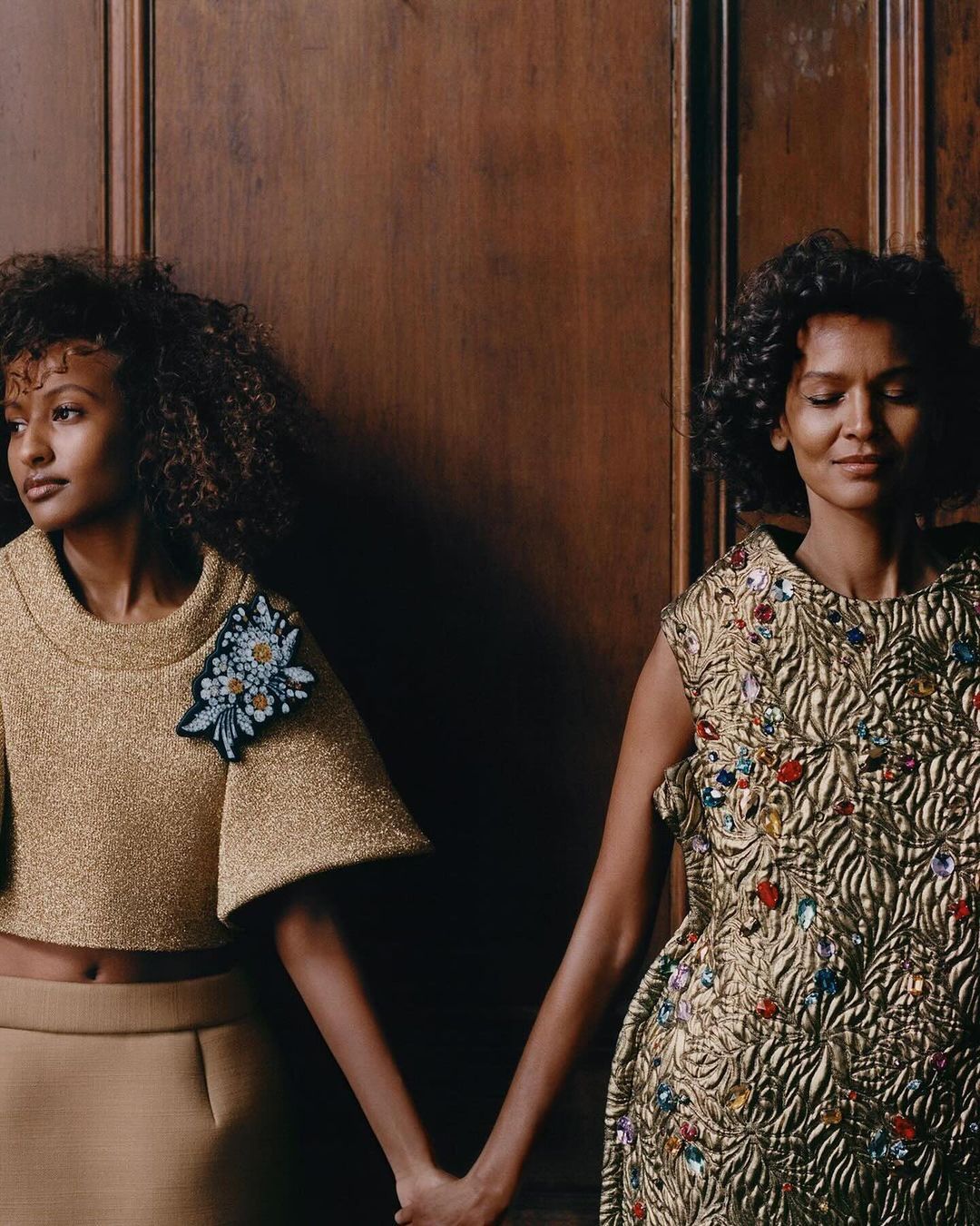Two women in embellished gold dresses hold hands against a dark wood wall in a studio portrait with medium framing and neutral colors.