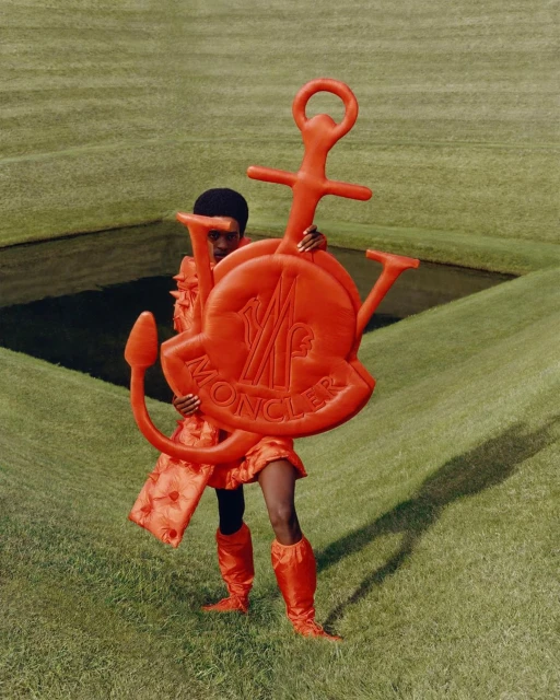 Fashion editorial shot of a Black woman in an orange outfit holding a large Moncler logo on a green lawn with square water features.