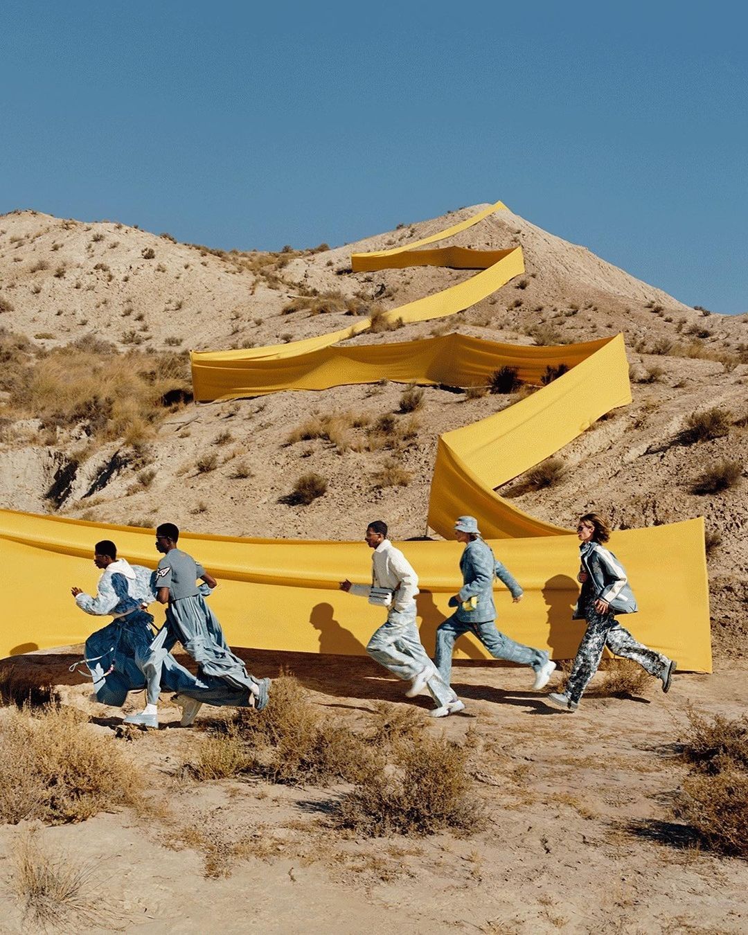 Five people run across a desert landscape with a long yellow banner winding up a sandy hill under a clear blue sky.