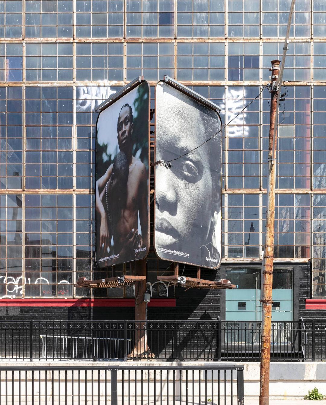 Two large black and white billboard portraits of a man and woman on a metal structure with a weathered industrial building behind.