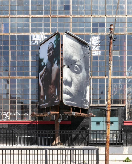 Two large black and white billboard portraits of a man and woman on a metal structure with a weathered industrial building behind.