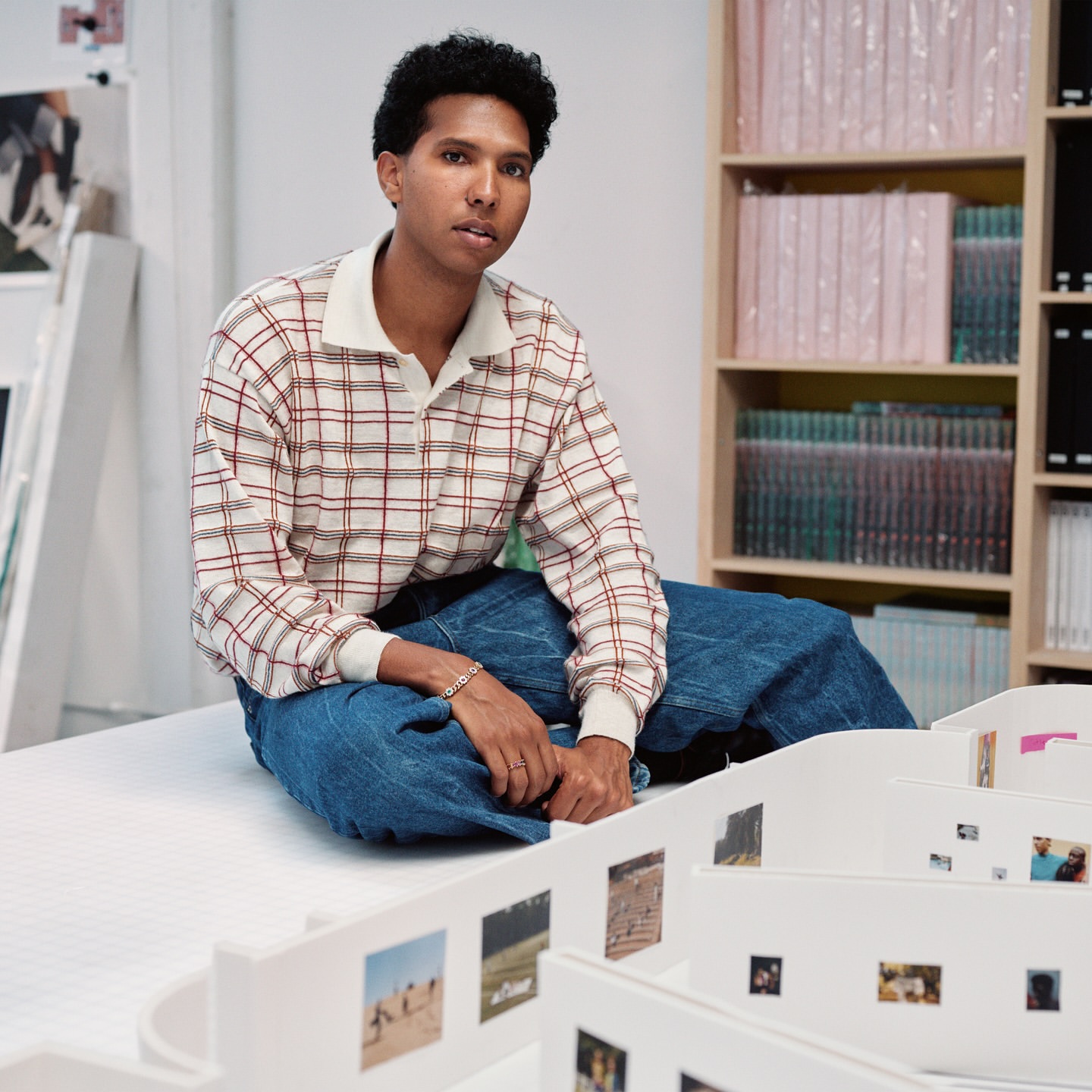 Adult wearing plaid shirt and blue jeans, seated indoors. White shelves with books and small printed photos in the background.