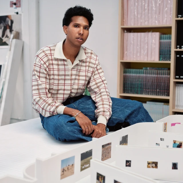 Adult wearing plaid shirt and blue jeans, seated indoors. White shelves with books and small printed photos in the background.
