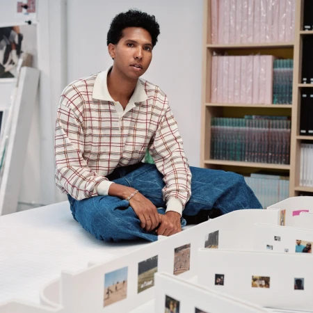 Adult wearing plaid shirt and blue jeans, seated indoors. White shelves with books and small printed photos in the background.