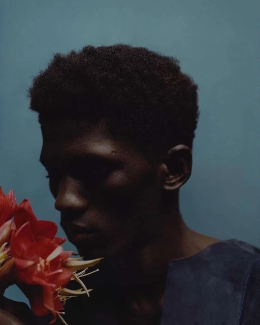 Portrait of a dark-skinned adult male in dark blue clothing with short natural hair holding a bouquet of amaryllis flowers against a teal background.