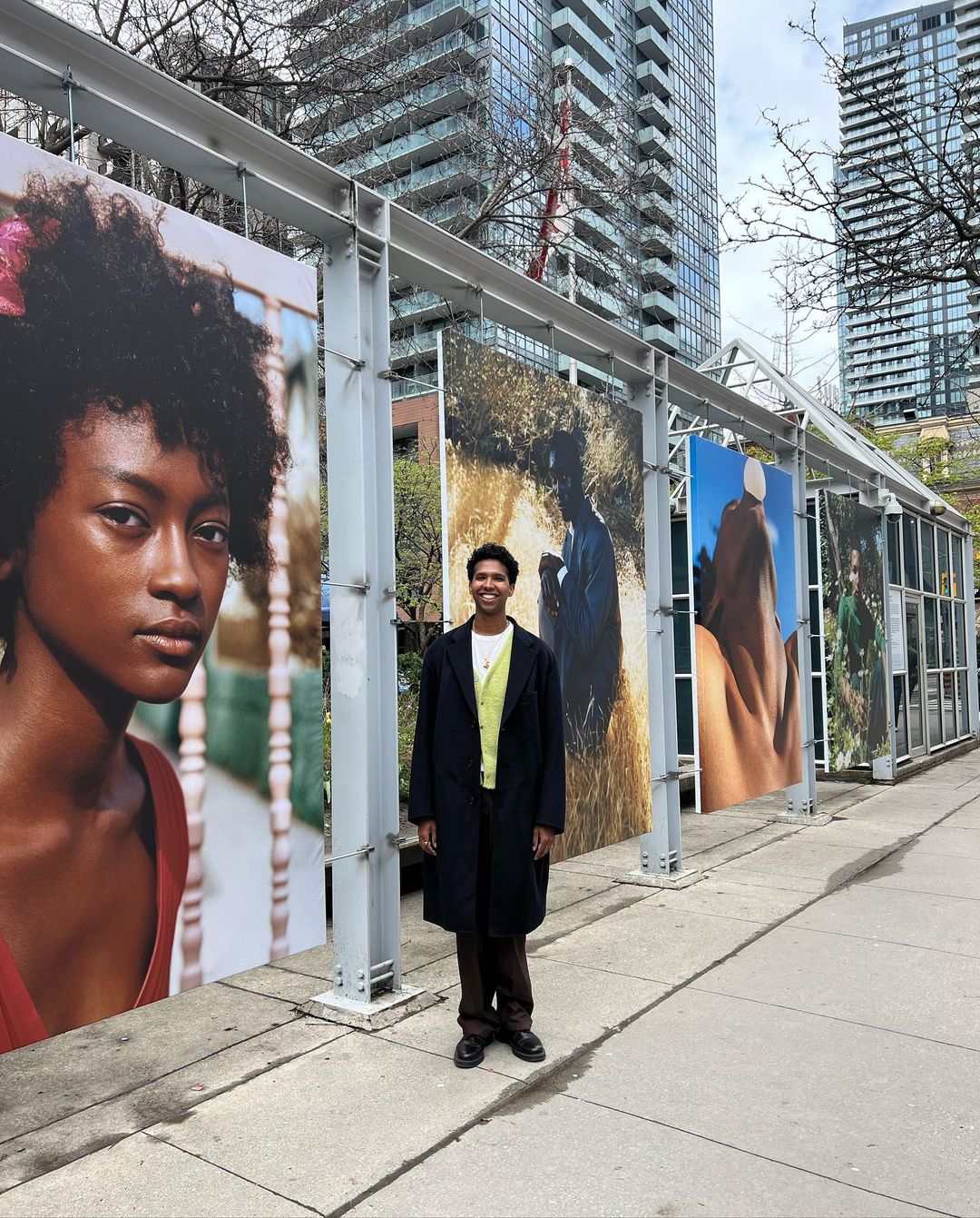 Young adult stands next to outdoor art exhibit of portrait photography in an urban park setting with modern skyscrapers in the distance.