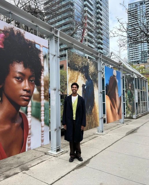 Young adult stands next to outdoor art exhibit of portrait photography in an urban park setting with modern skyscrapers in the distance.