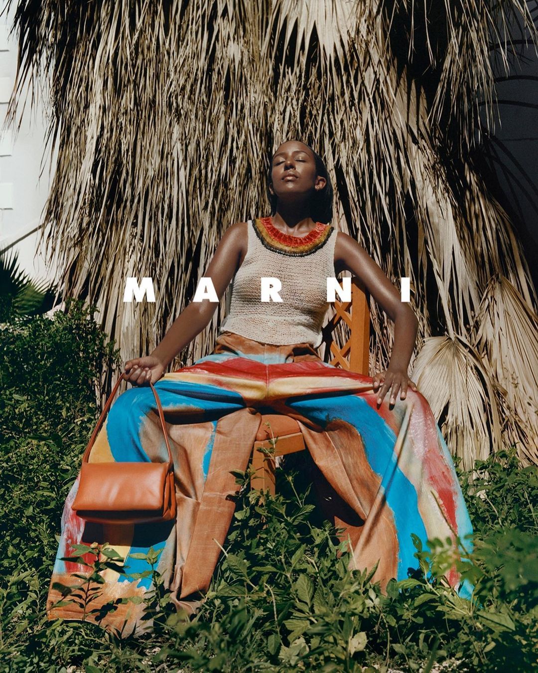 Woman in patterned pants sits in greenery outdoors, holding orange bag. The scene features lush vegetation and sunlight.