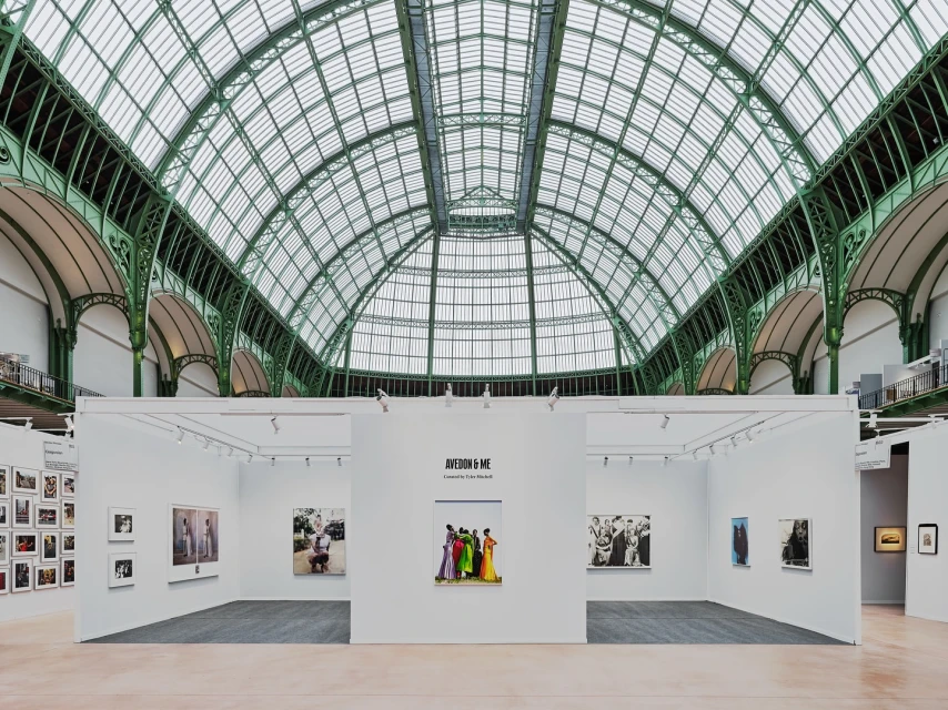 Interior view of an art exhibition featuring photograph displays within a hall with a large arched, green-framed glass ceiling.