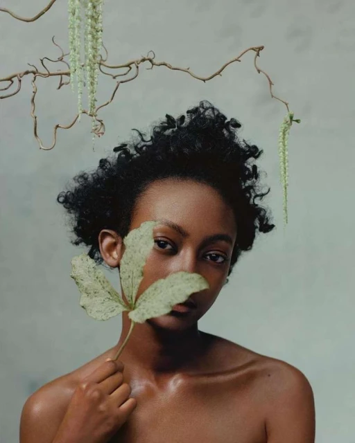 Striking portrait of a young Black woman with a leaf held to her face, set against a muted grey background with botanical elements.