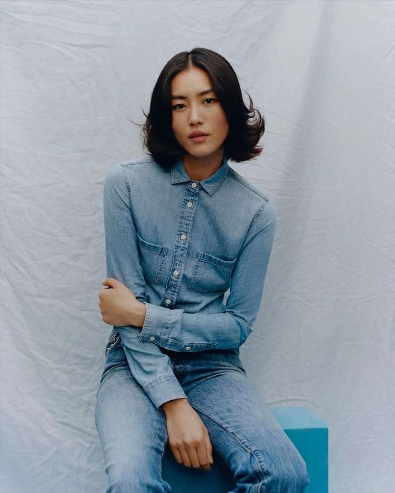 Young woman in denim shirt and jeans sitting in a photo studio against a textured background with neutral lighting.