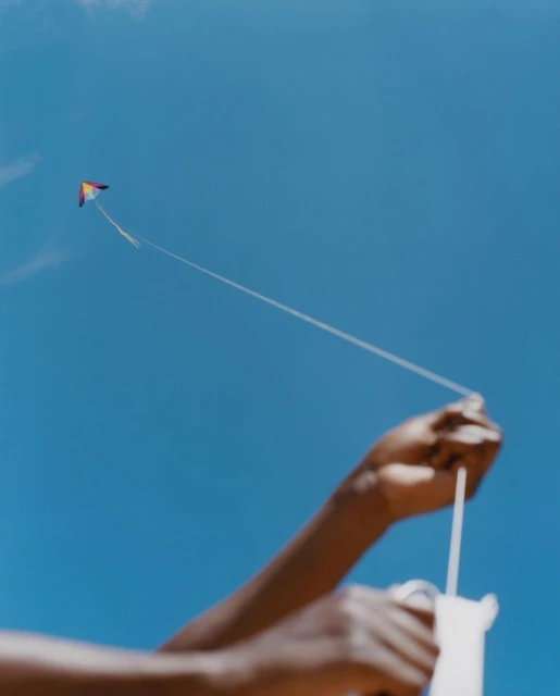 Person holding a kite string with a colorful kite flying in a clear blue sky during the day in a low-angle outdoor shot.