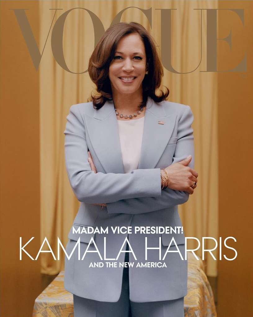 Kamala Harris poses on Vogue cover in a light blue blazer and pantsuit with a soft smile. She is standing in front of a gold curtain.