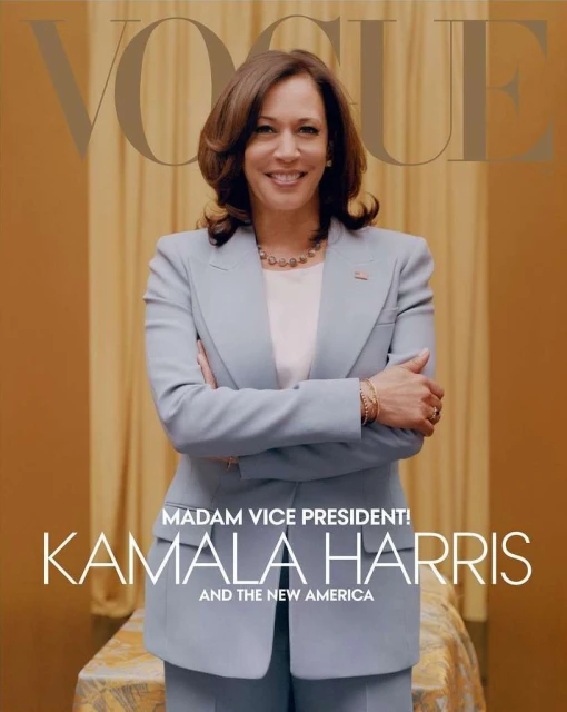 Kamala Harris poses on Vogue cover in a light blue blazer and pantsuit with a soft smile. She is standing in front of a gold curtain.