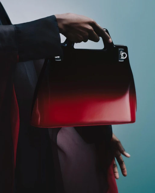 Close-up of a Black person holding a red-to-black ombre leather purse with silver hardware against a teal background in a fashion shot.