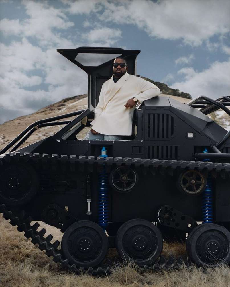 Man wearing a white coat poses in a black tank-like vehicle in a dry grassy field under a cloudy sky. Fashion editorial shot.
