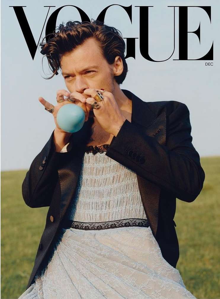 Vogue cover featuring Harry Styles in a light blue dress and black blazer, blowing up a blue balloon, in a grassy outdoor setting.