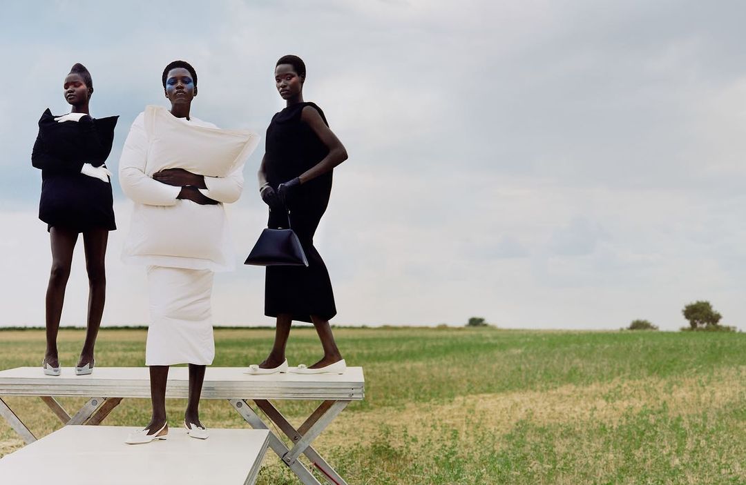 Three dark-skinned fashion models in unique black-and-white dresses pose on a white platform in a grassy field under a cloudy sky.