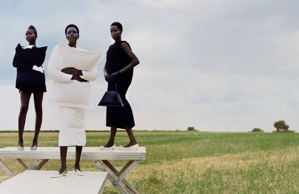 Three dark-skinned fashion models in unique black-and-white dresses pose on a white platform in a grassy field under a cloudy sky.