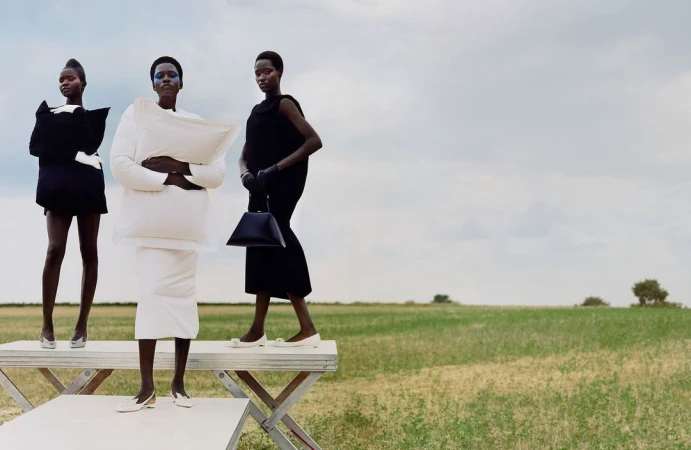 Three dark-skinned fashion models in unique black-and-white dresses pose on a white platform in a grassy field under a cloudy sky.