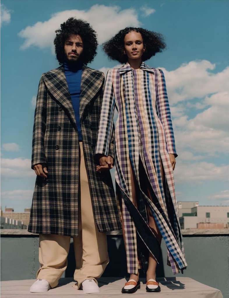 A stylish couple, a man and a woman, stand close on a rooftop with city views. They wear plaid coats and hold hands, under a cloudy sky.