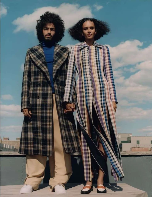 A stylish couple, a man and a woman, stand close on a rooftop with city views. They wear plaid coats and hold hands, under a cloudy sky.