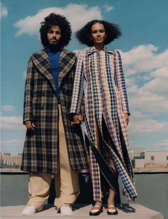A stylish couple, a man and a woman, stand close on a rooftop with city views. They wear plaid coats and hold hands, under a cloudy sky.