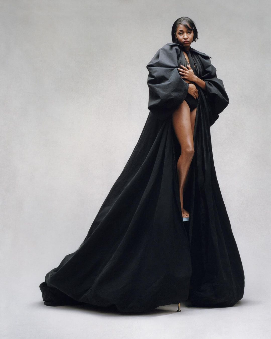 A woman with short dark hair is standing in a photo studio wearing a black bodysuit and a black puffy robe with clear high heels.