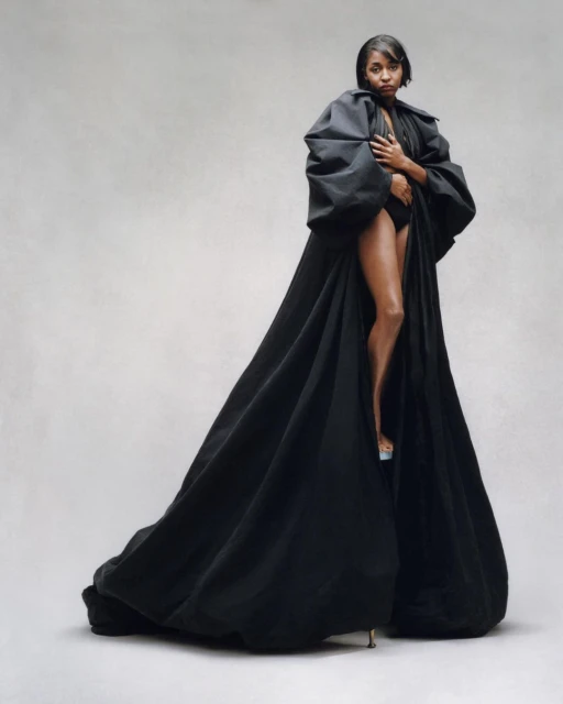 A woman with short dark hair is standing in a photo studio wearing a black bodysuit and a black puffy robe with clear high heels.