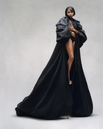 A woman with short dark hair is standing in a photo studio wearing a black bodysuit and a black puffy robe with clear high heels.