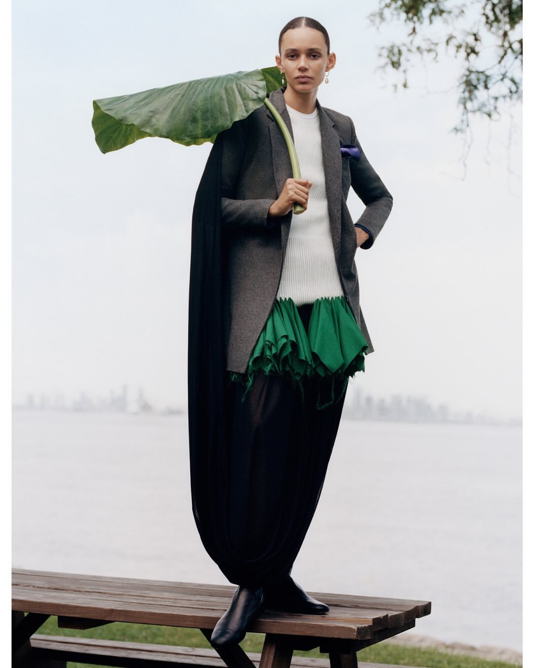 Model standing outdoors on a wooden bench holding a large green leaf over their shoulder, wearing a blazer and dark trousers.