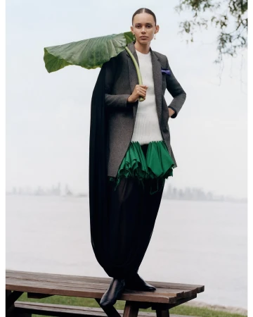 Model standing outdoors on a wooden bench holding a large green leaf over their shoulder, wearing a blazer and dark trousers.