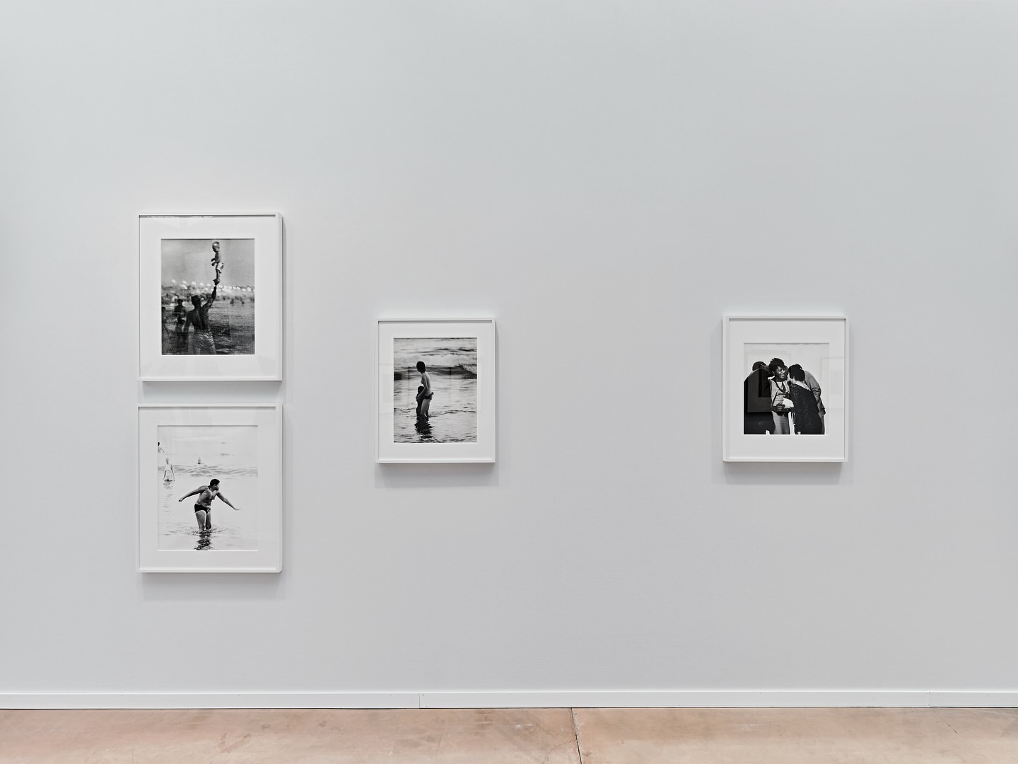 Black and white photographs of people at the beach hang in white frames on a white wall, above a light brown floor, in a gallery.