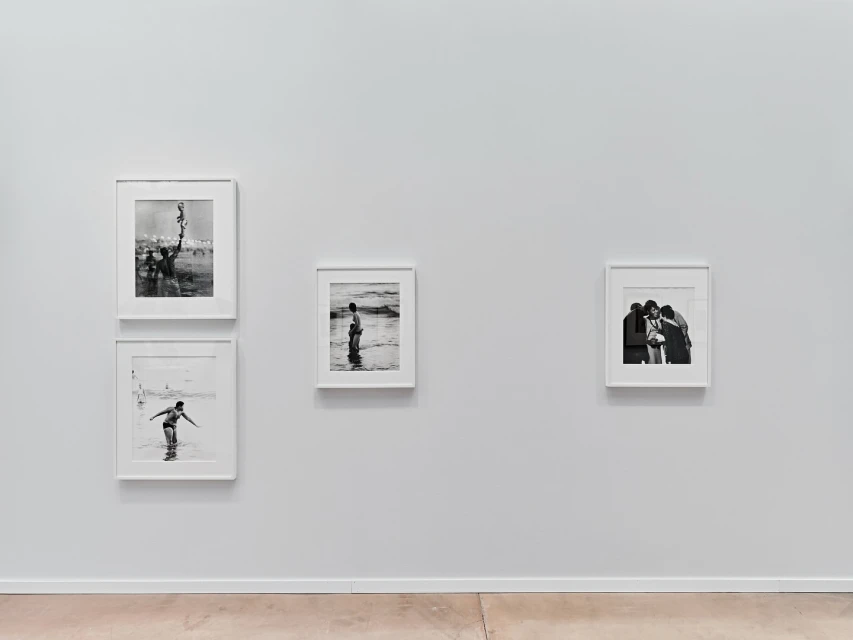 Black and white photographs of people at the beach hang in white frames on a white wall, above a light brown floor, in a gallery.