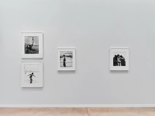 Black and white photographs of people at the beach hang in white frames on a white wall, above a light brown floor, in a gallery.