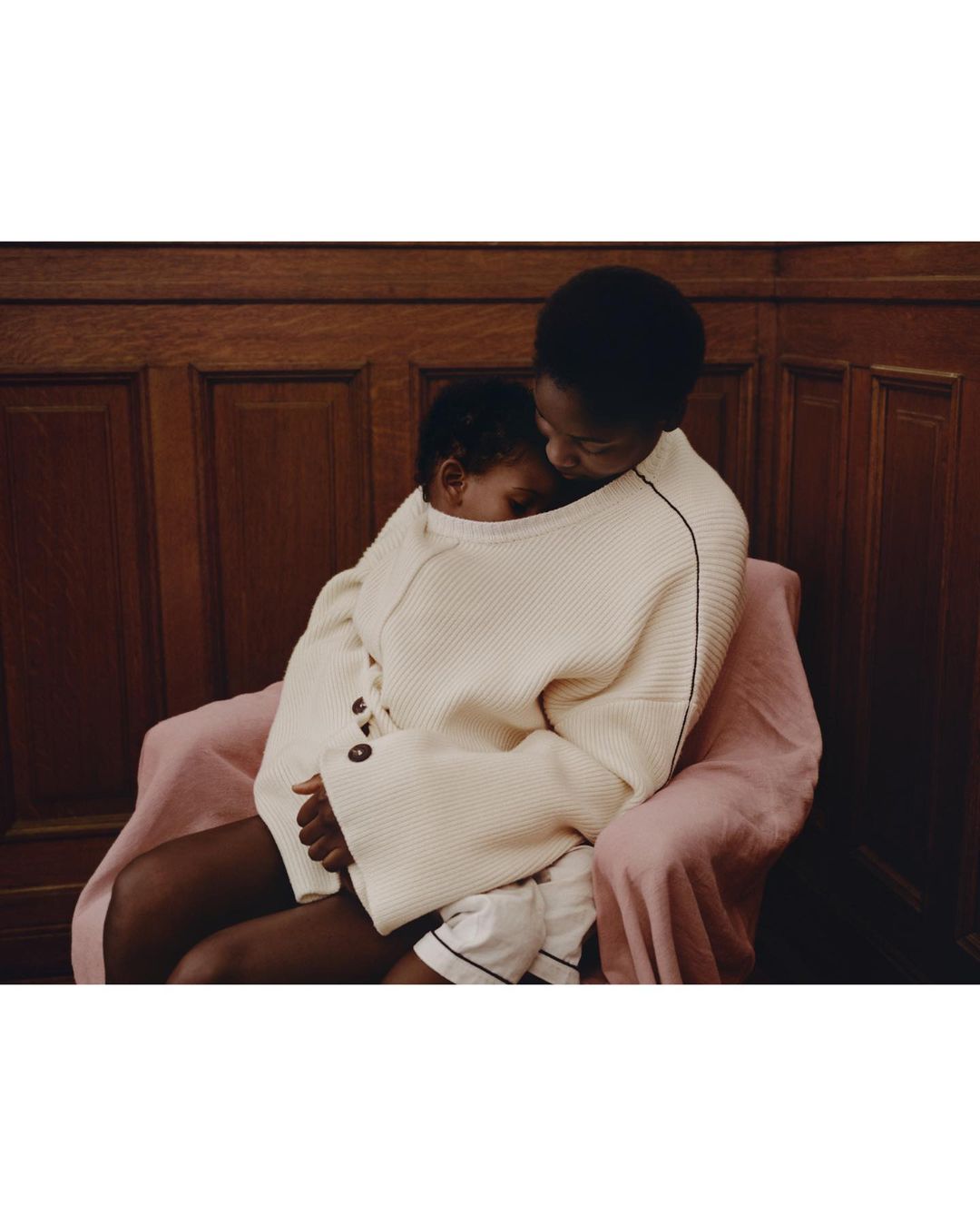 Dark-skinned woman in a cream waffle knit sweater hugs a baby wearing matching cream knit. Pink blanket, wood paneling in background.