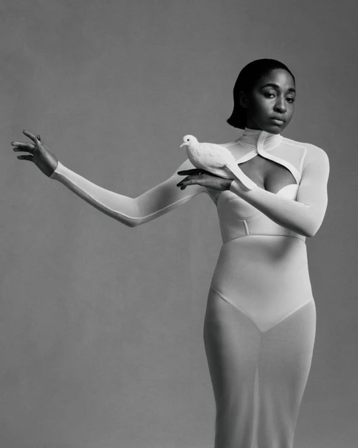 Elegant studio portrait of an African American woman in a white gown, holding a white dove, in a monochrome setting with soft lighting.