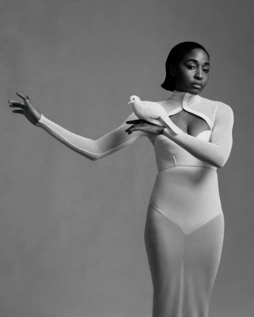 Elegant studio portrait of an African American woman in a white gown, holding a white dove, in a monochrome setting with soft lighting.