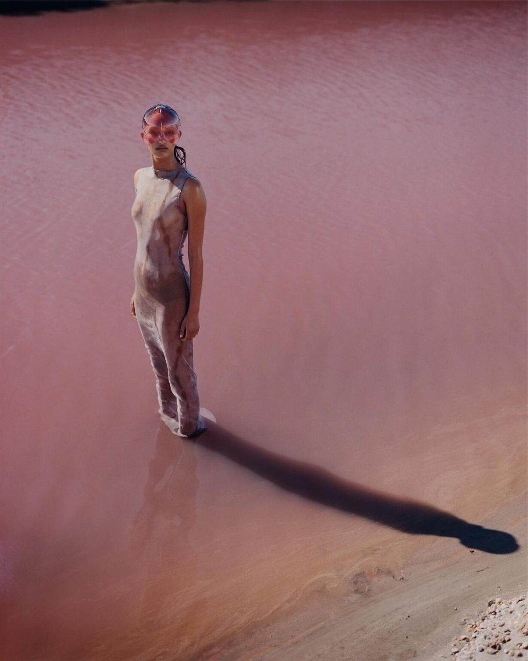 Artistic fashion shot featuring a woman in a pale dress standing in pink water, with a long shadow and surreal makeup and headpiece.