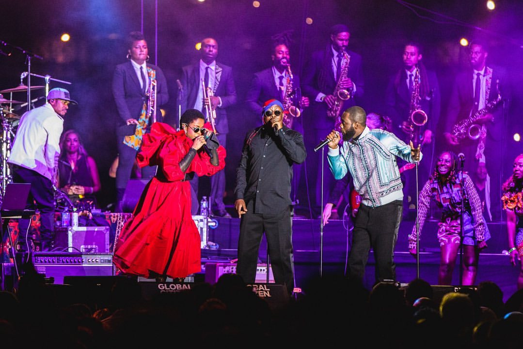 Live concert stage with three singers in the foreground performing in front of a band with saxophones under purple stage lighting.